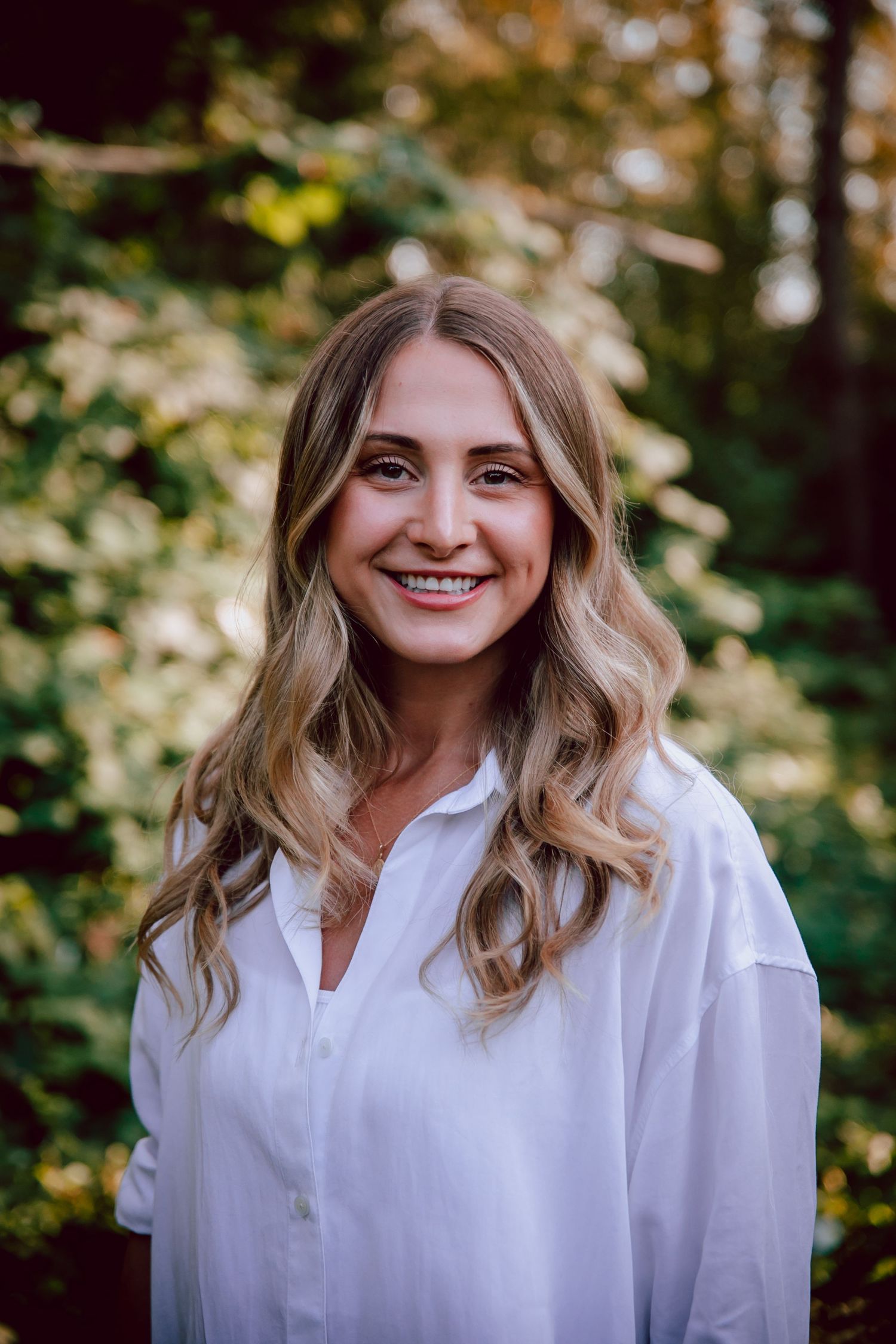 Outdoor portrait of woman with blonde wavy hair wearing white shirt against nature backdrop.