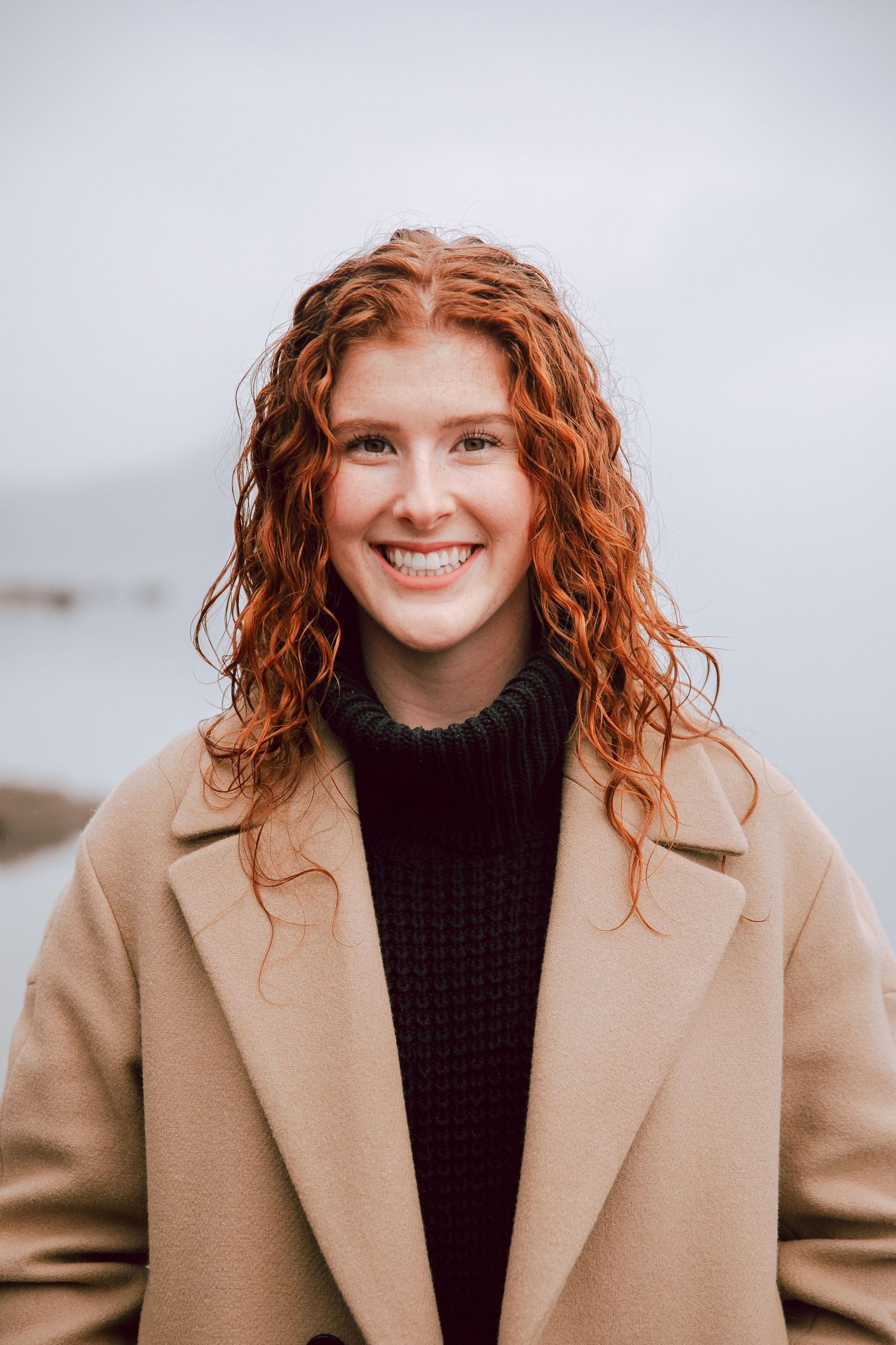 Portrait of woman with curly red hair wearing beige coat and black sweater against grey background.