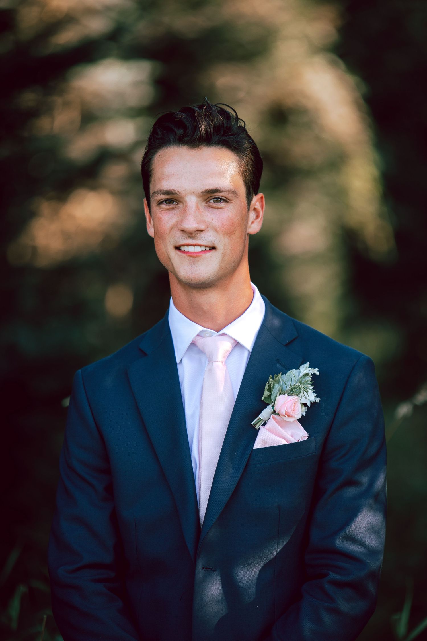 A groom in a navy blue suit with a pink tie and boutonniere poses for a formal portrait outdoors.