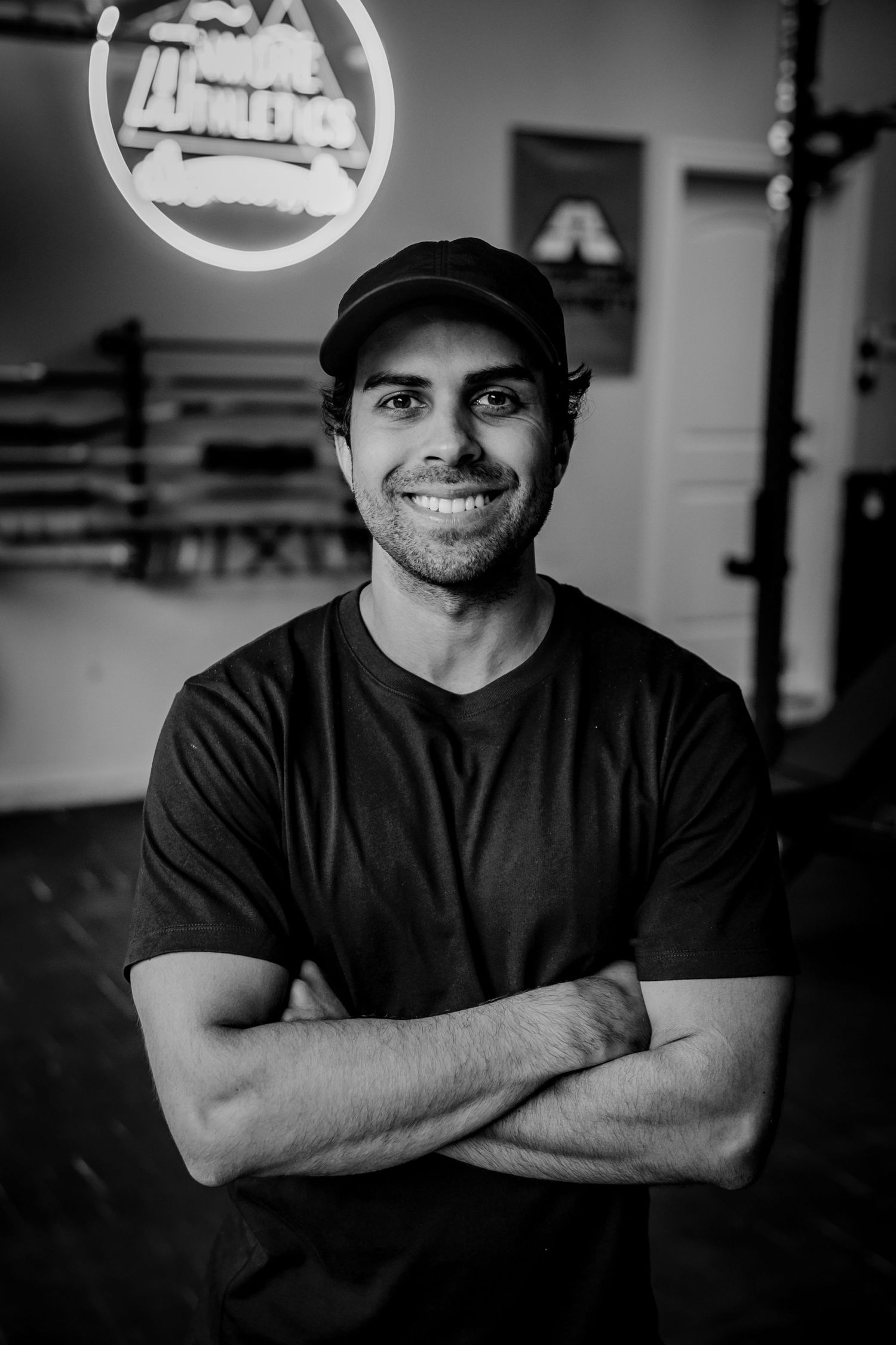 Man in black t-shirt and cap smiling in front of illuminated business sign.