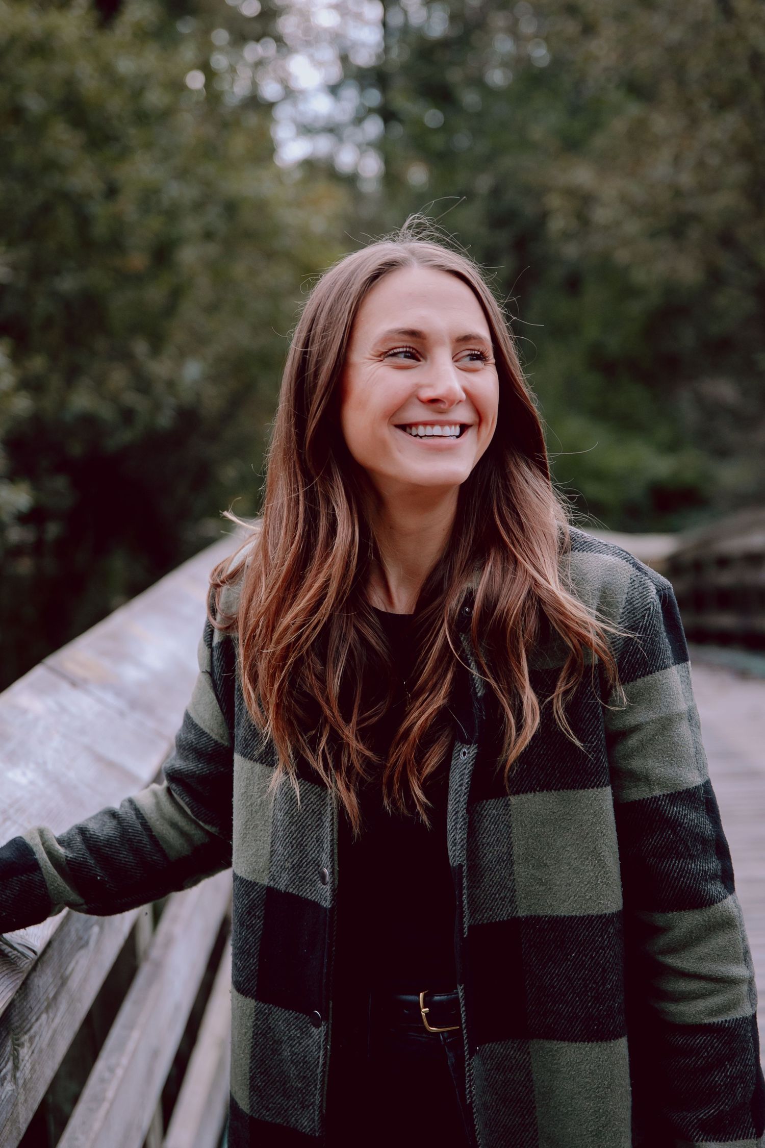 Woman in green plaid jacket smiling while standing on wooden bridge outdoors.