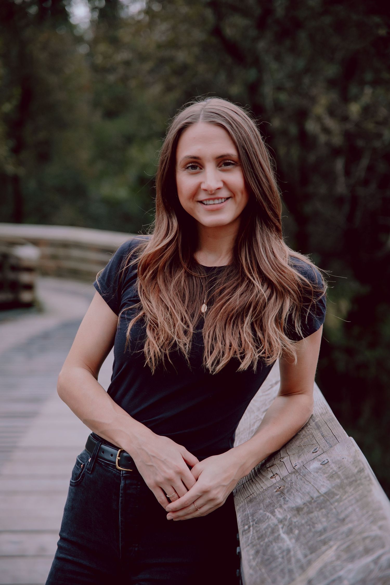 Woman in black shirt smiling while leaning against wooden railing on outdoor path.