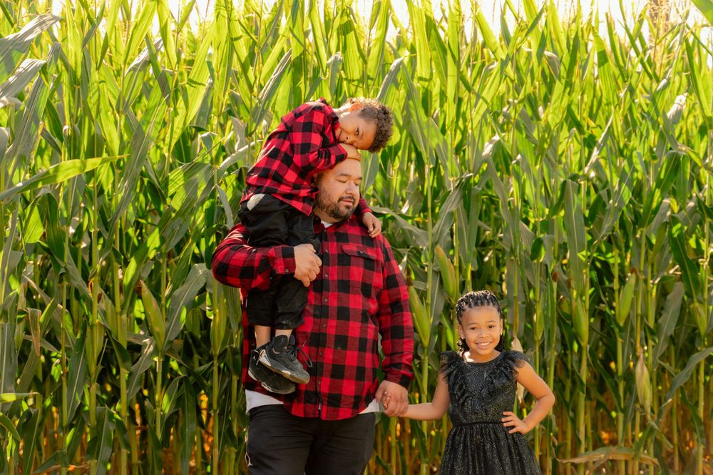 Two people in red plaid flannel and black outfits pose playfully in front of tall corn stalks.