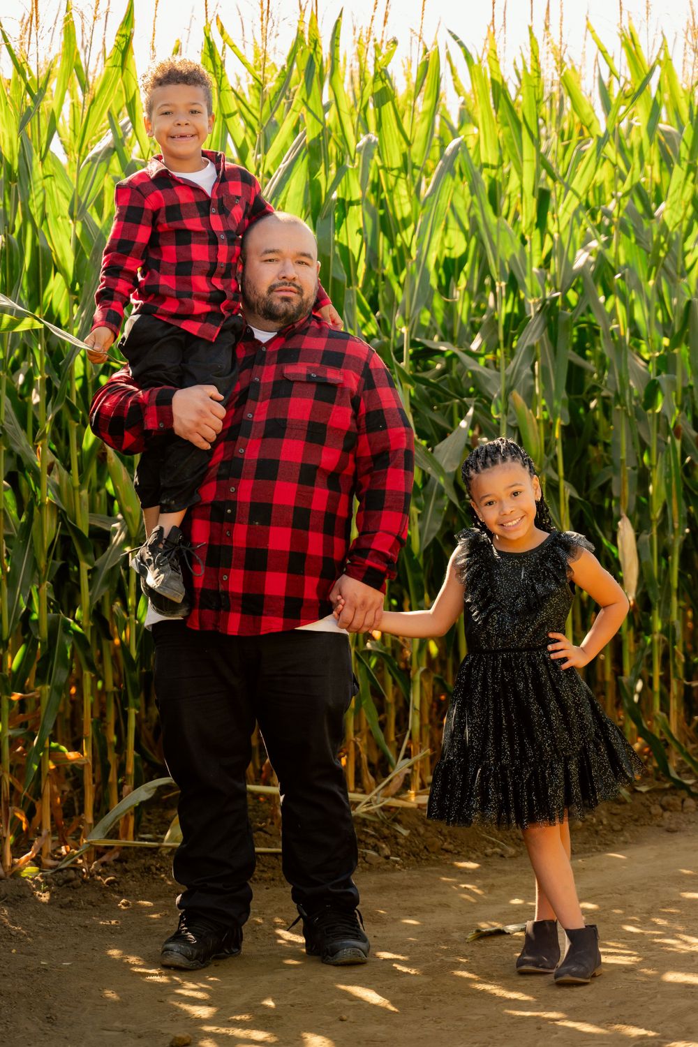 A pair in coordinating red buffalo plaid and black outfits stand together by a cornfield.
