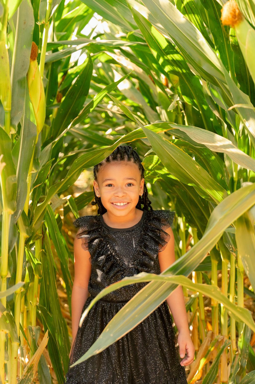 A young person in a black dress smiles brightly while standing in a corn field.