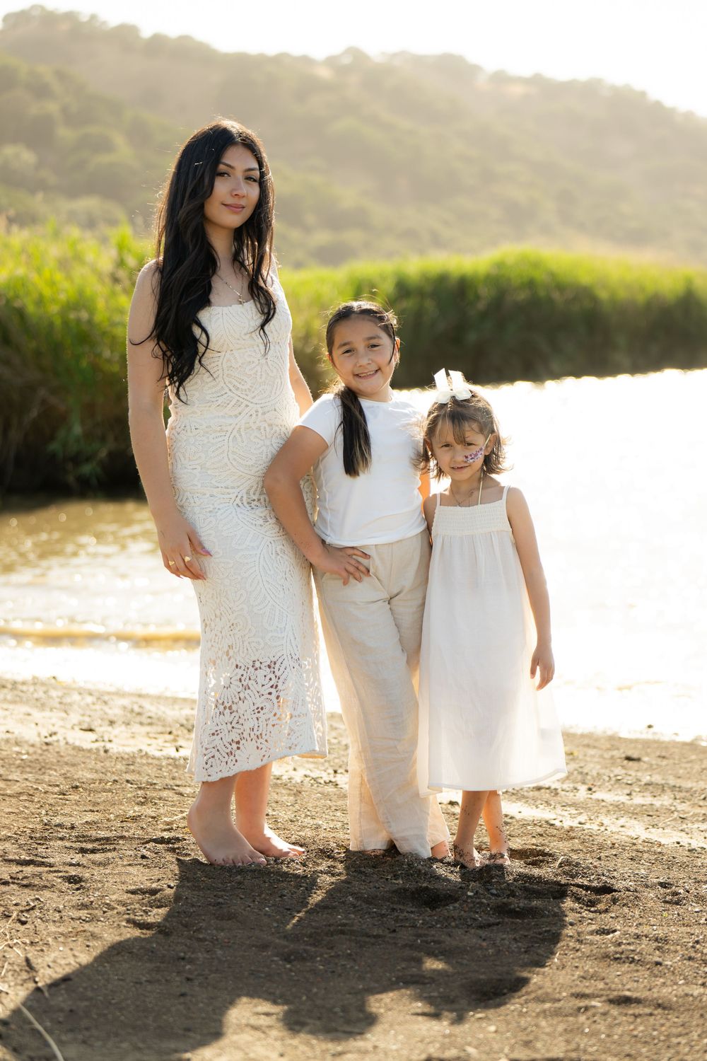 A sweet beach photoshoot featuring white lace and flowy dresses against a scenic coastal backdrop at sunset.