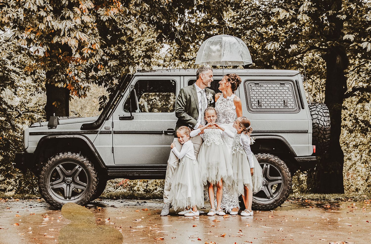 Group in white outfits posing in front of grey SUV in rainy weather. BruidsfotoNederland.