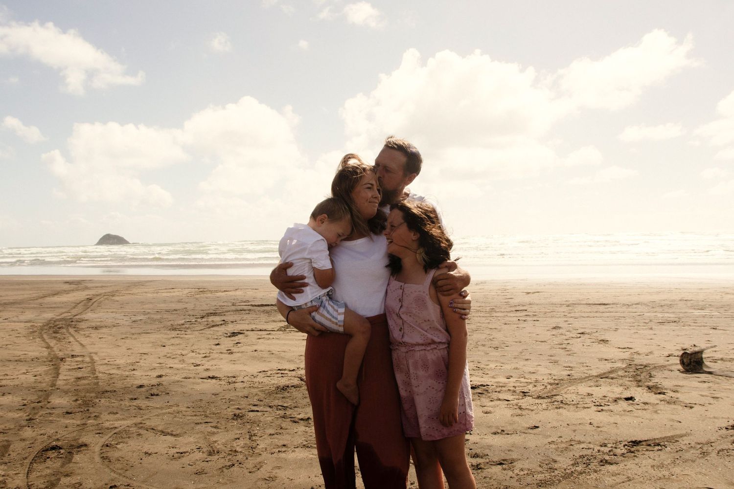 A family of four, mum, dad, little brother and big sister, at the beach at golden hour. Dad has has his arms wrapped around his family