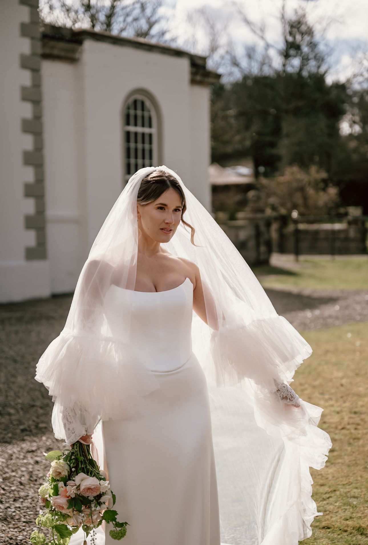 A bride poses for a portrait in front of the stunning Holesfoot in Cumbria. The golden glow of the sunshine accentuates her opulent veil.