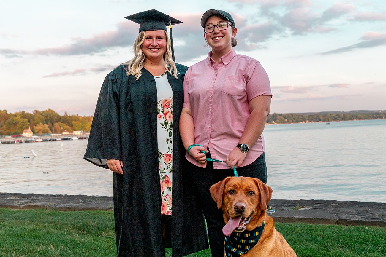 A graduation photo featuring a graduate in cap and gown alongside a companion and their brown dog by a waterfront at sunset.