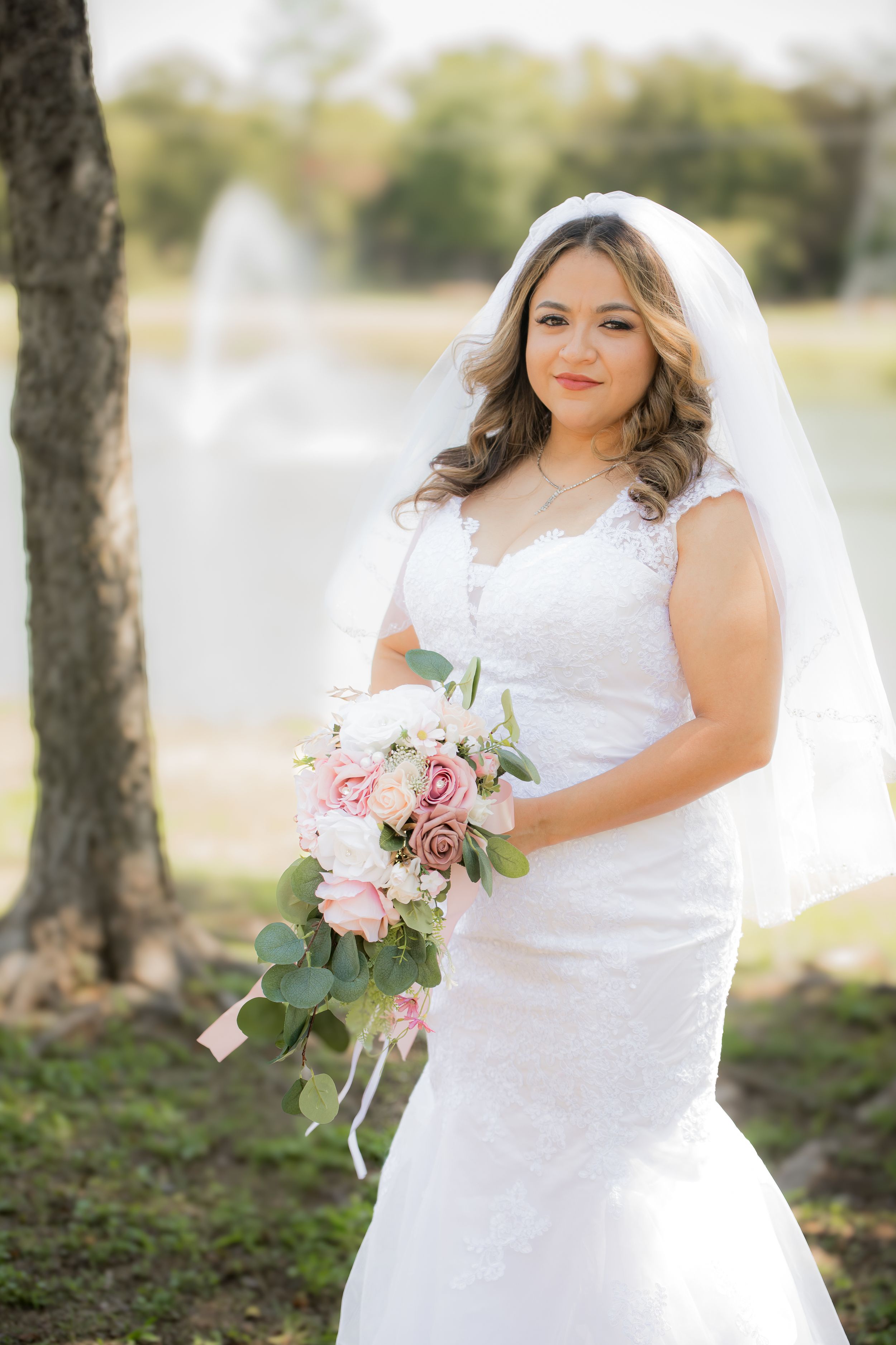 A bride in a lace mermaid wedding dress holds a pink and white bouquet outdoors.