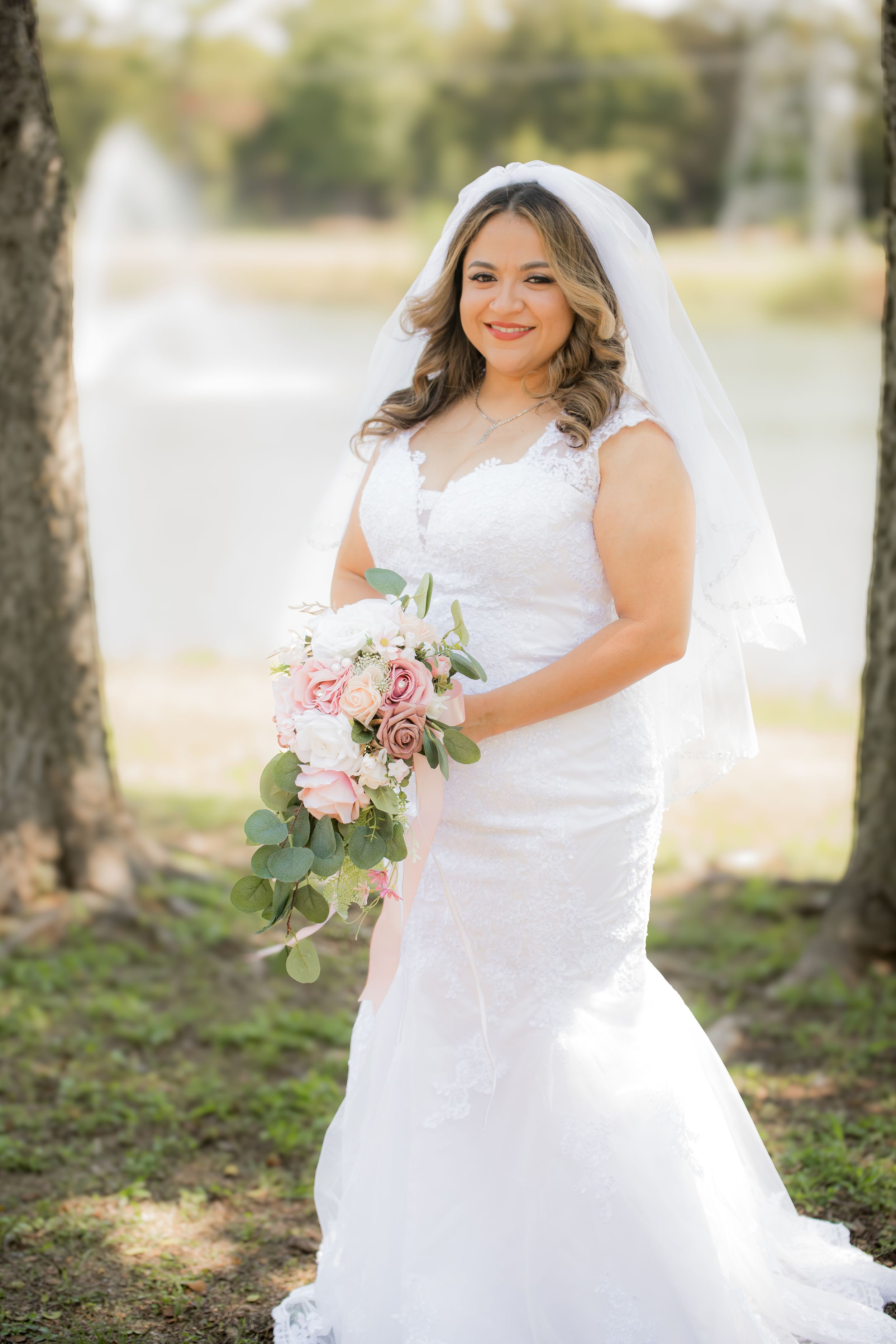 A bride poses with her bouquet of pink roses and eucalyptus in an outdoor setting.