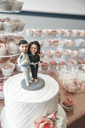 White wedding cake topped with a cute bride and groom figurine, surrounded by cupcakes.