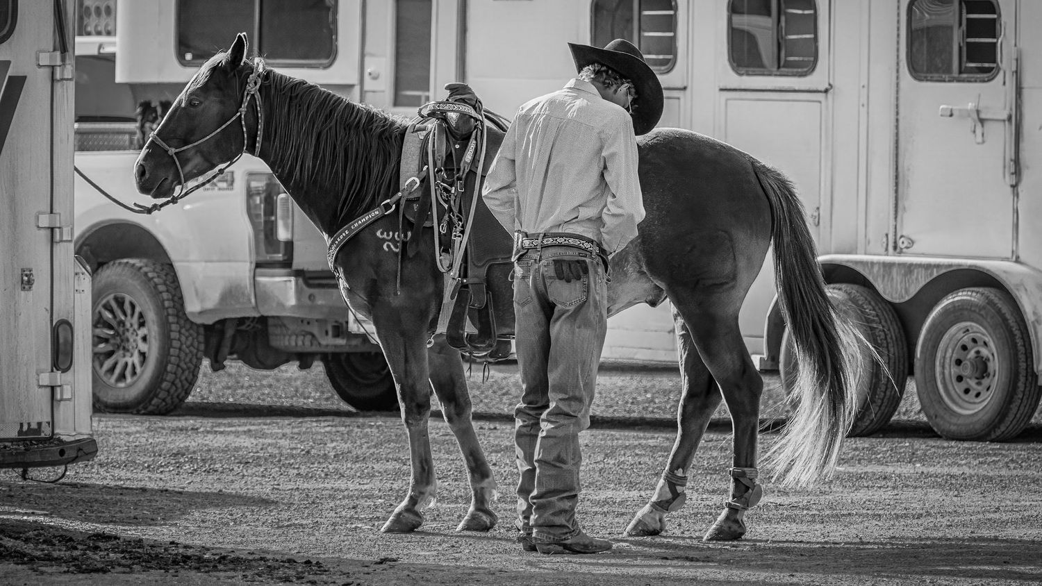 A black and white image showing someone with a horse next to a horse trailer in a rustic setting.
