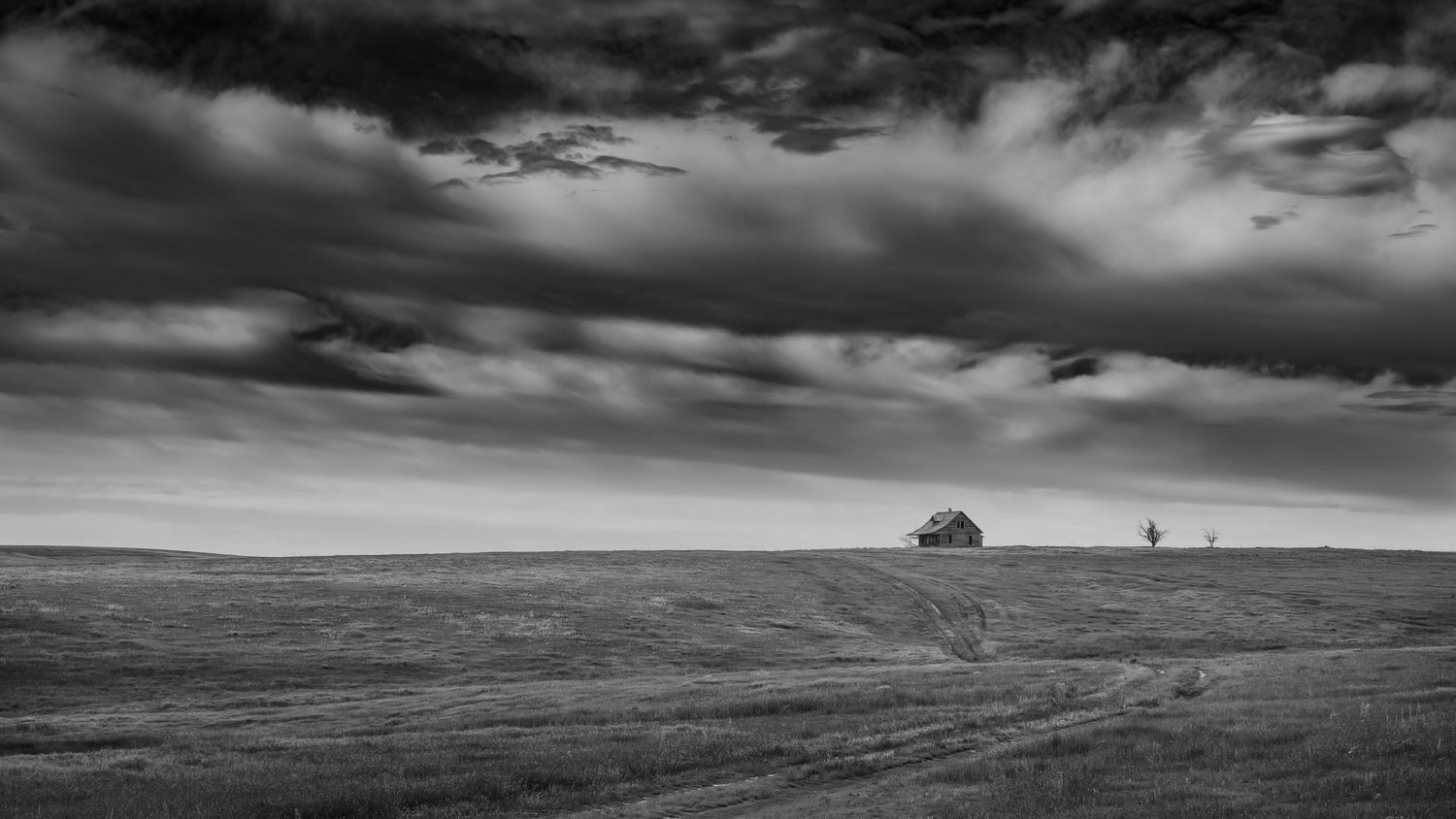 A dramatic black and white rural landscape with moody clouds above rolling fields and distant trees on the horizon.