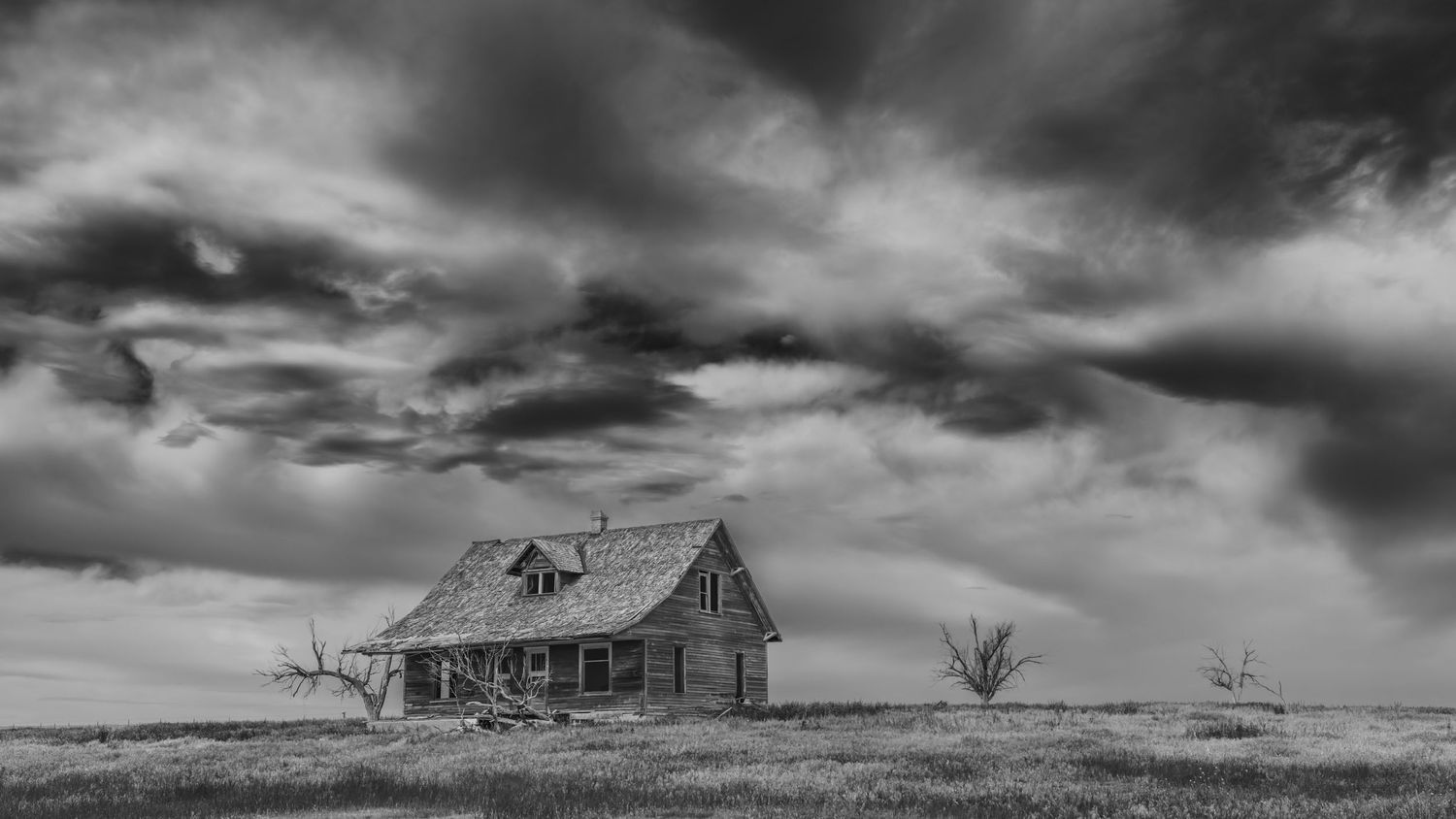 A dramatic black and white photograph of an abandoned farmhouse standing alone on a desolate prairie under stormy skies.