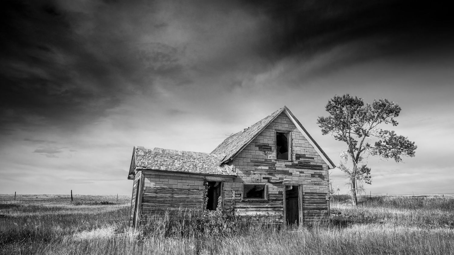 A dramatic black and white photo of an abandoned weathered farmhouse on an empty prairie.