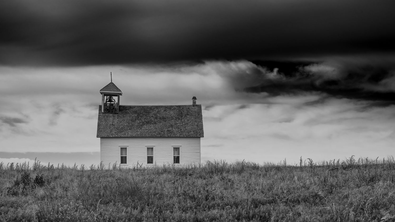 A black and white photograph of an abandoned rural church with a bell tower stands isolated in a grassy field.