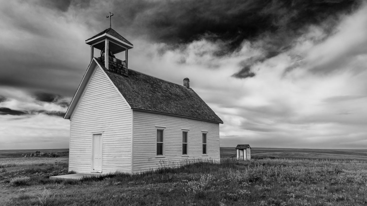 A stark black and white photograph of a white wooden country church under dramatic storm clouds.