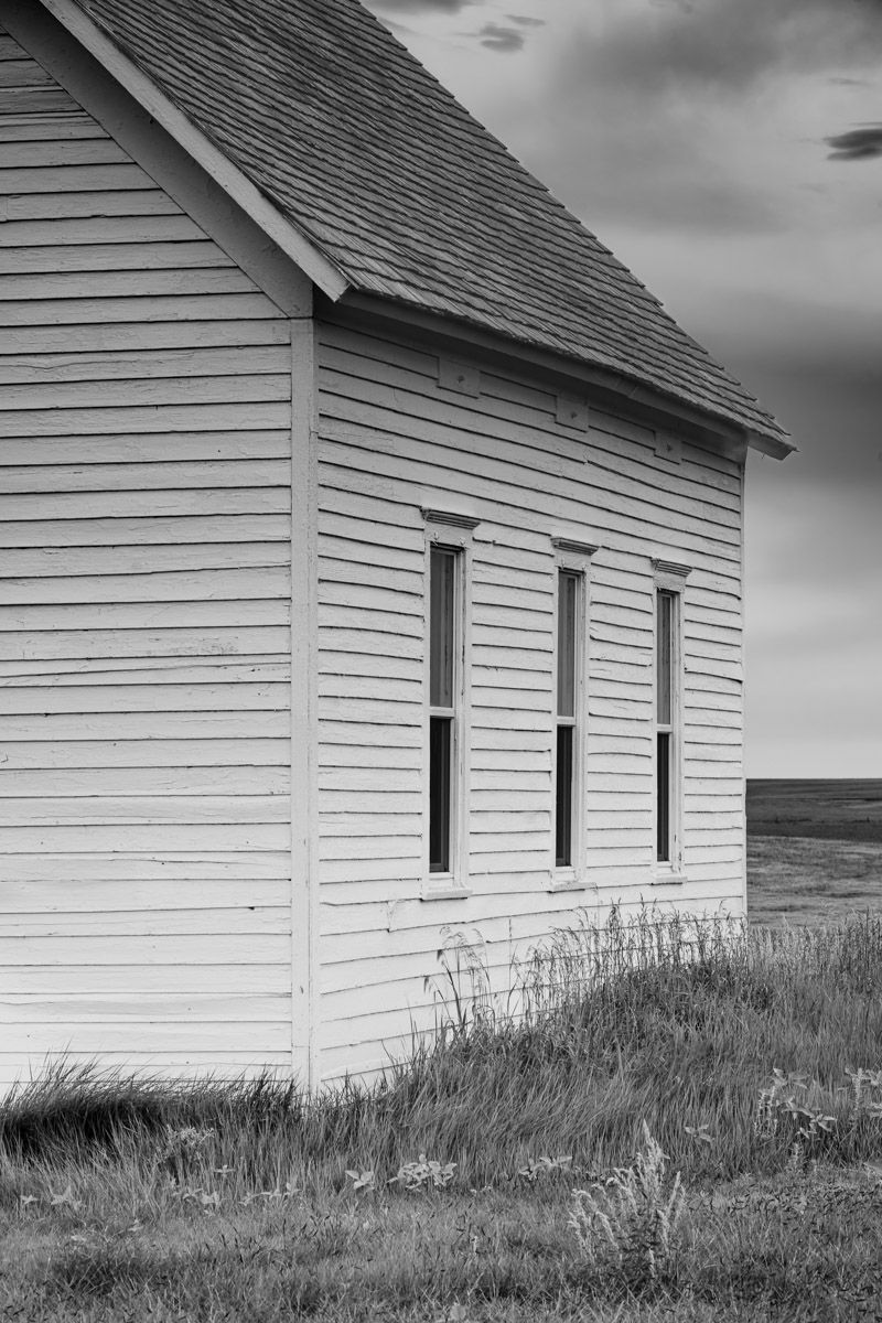 Black and white image of a rural white clapboard church with three windows against a prairie landscape.
