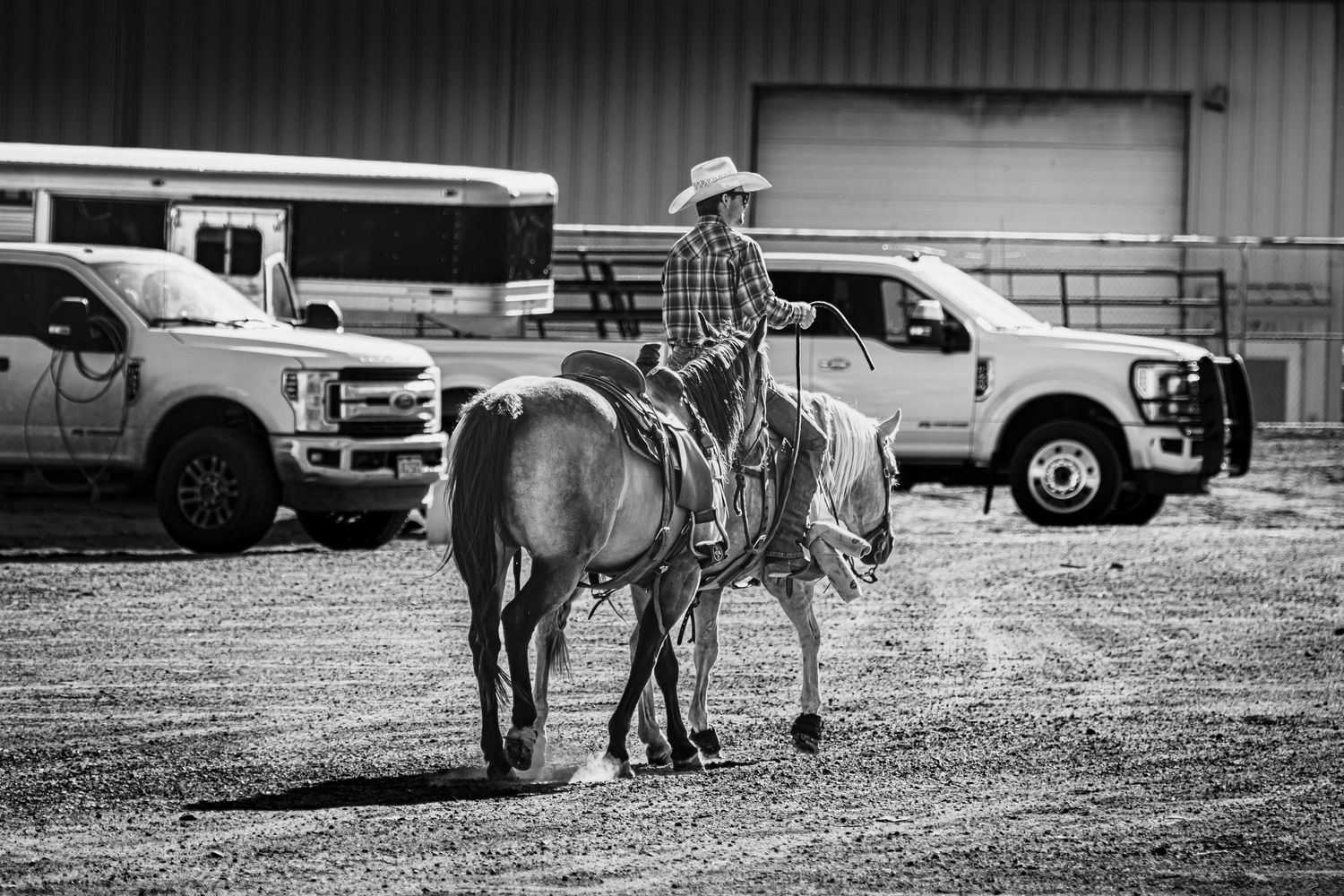 Cowboy seated on horseback in black and white photo with pickup trucks parked in the background.