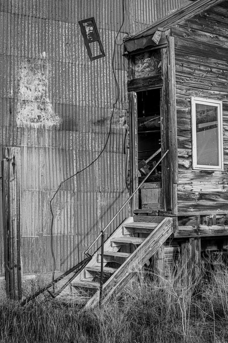 Black and white photo of weathered wooden stairs leading to doorway of abandoned building with corrugated metal wall.