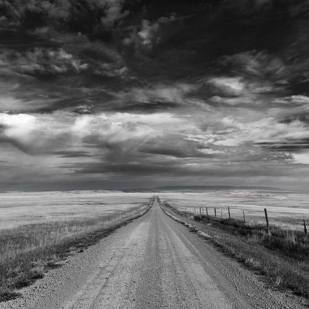 A dramatic black and white country dirt road stretches to the horizon under stormy prairie skies with fence posts.