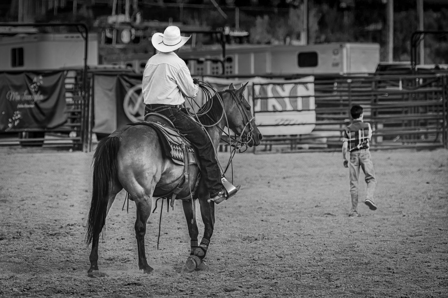 A cowboy rides a dark horse in an arena during a rodeo event while a figure stands nearby in black and white photography.
