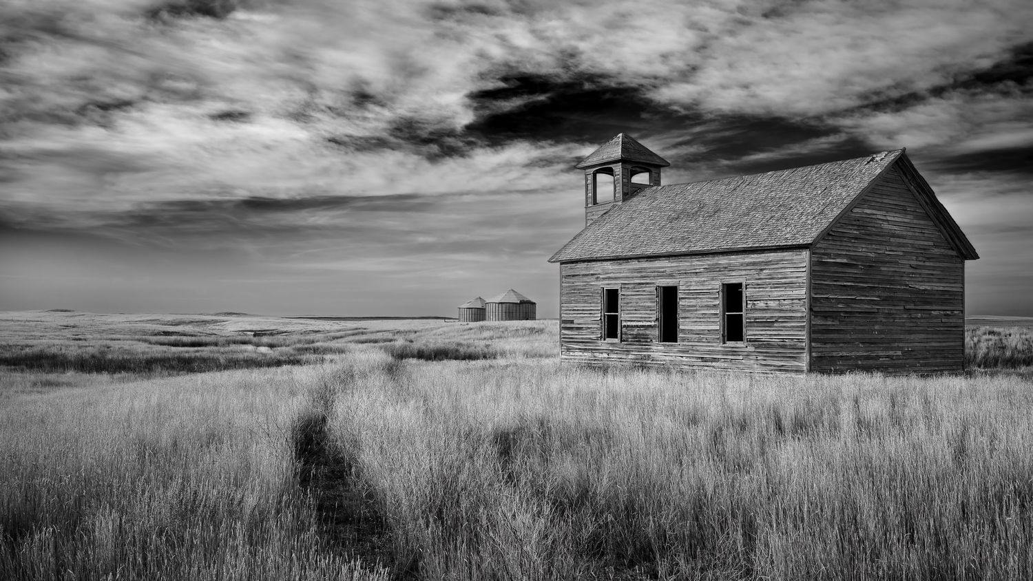 Abandoned wooden church stands alone in a grassy prairie field under dramatic clouds in black and white.