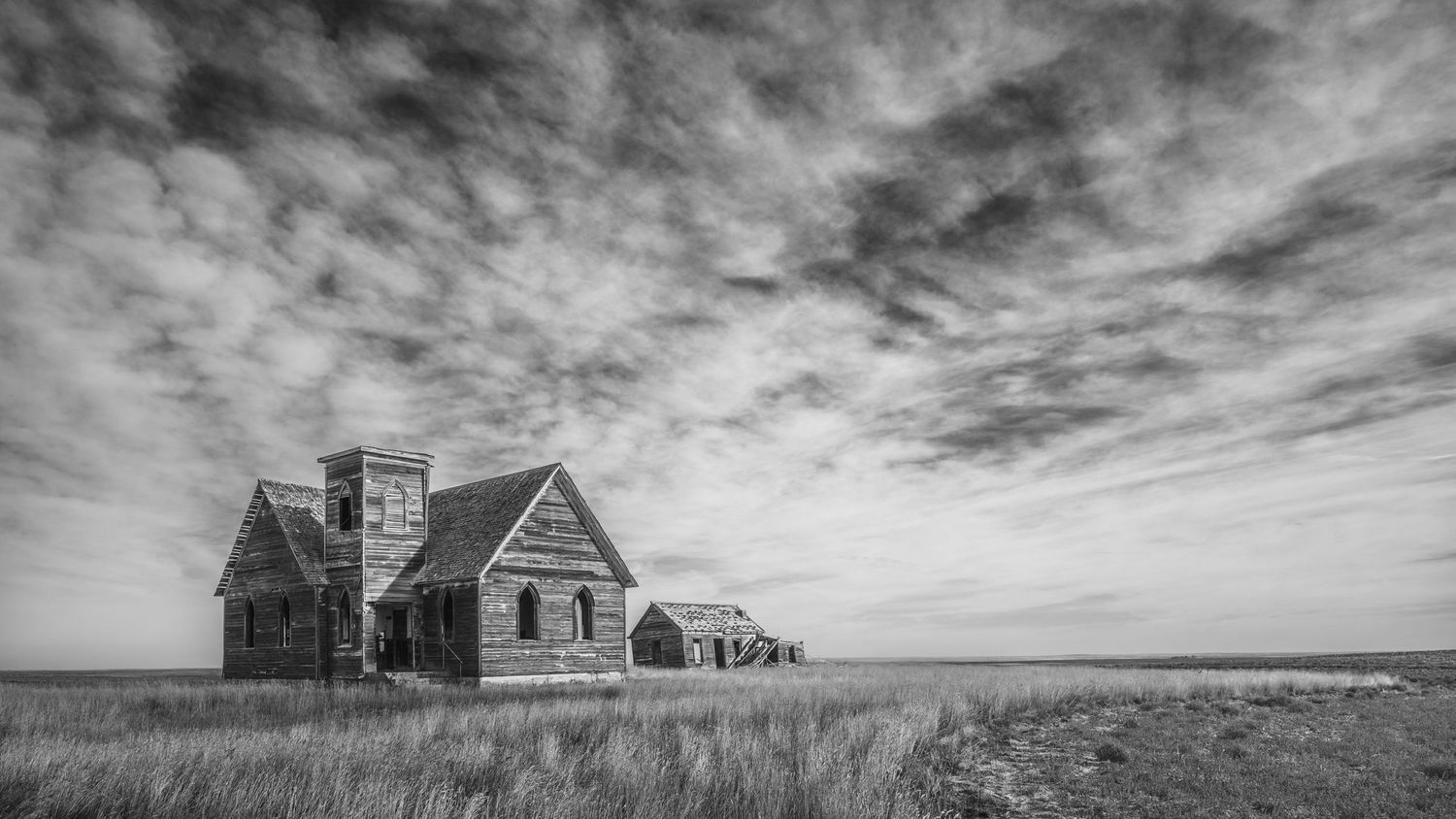 An abandoned farmhouse stands alone on the prairie under dramatic cloudy skies in black and white.