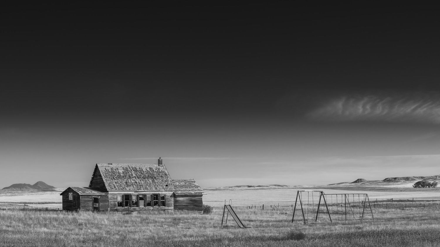 An abandoned old barn and empty swing set stand in a desolate prairie landscape under dramatic black and white skies.