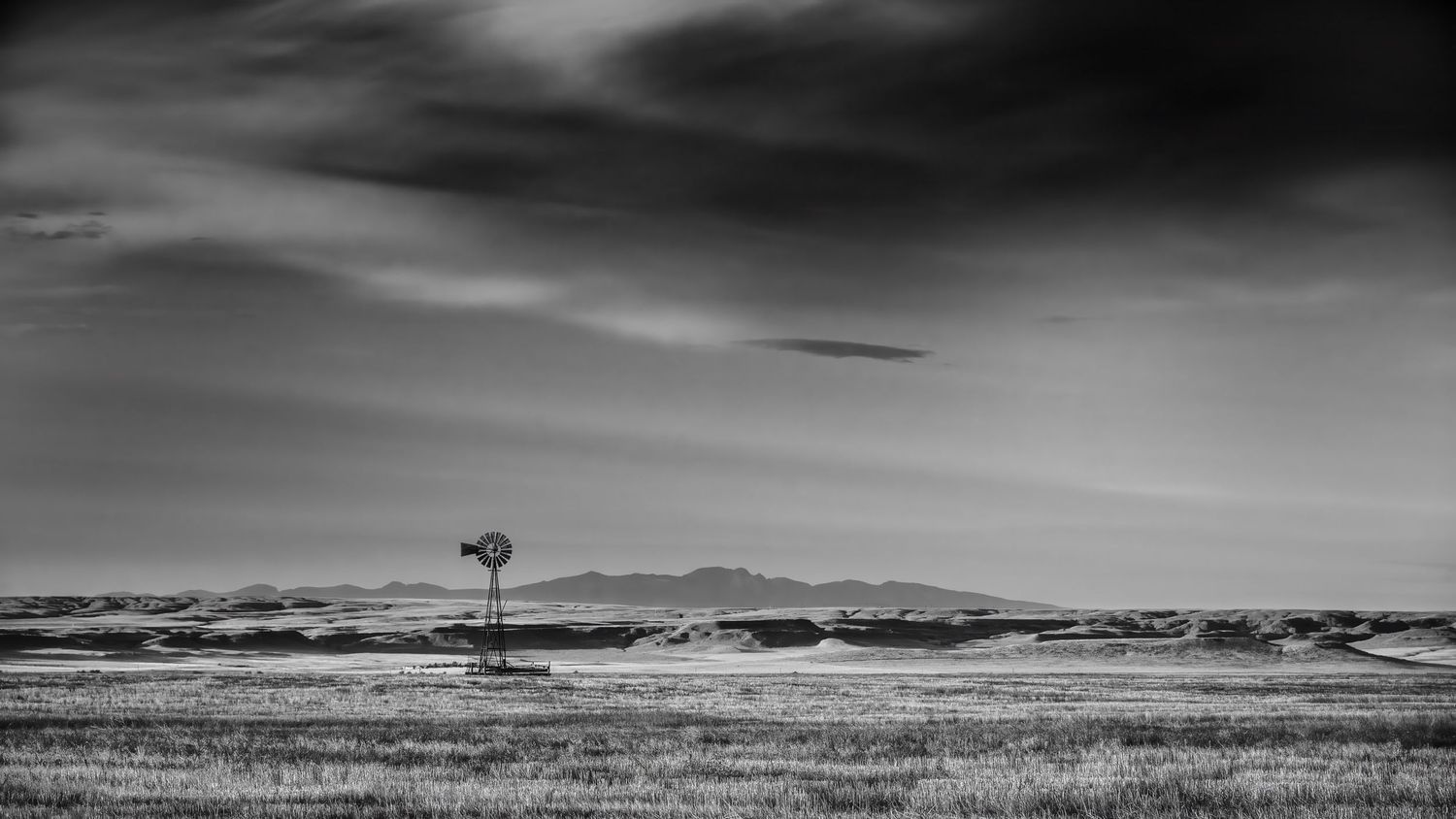 A lone windmill stands in a vast prairie landscape under dramatic black and white skies with mountains in the distance.