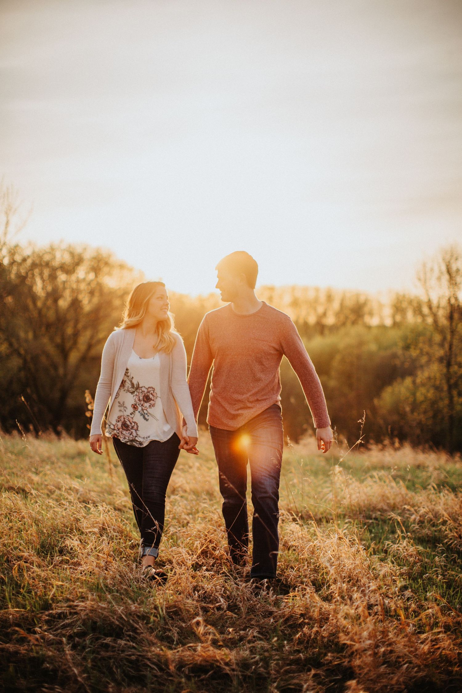 A couple walks hand in hand through a golden field at sunset with warm backlighting creating a romantic silhouette.