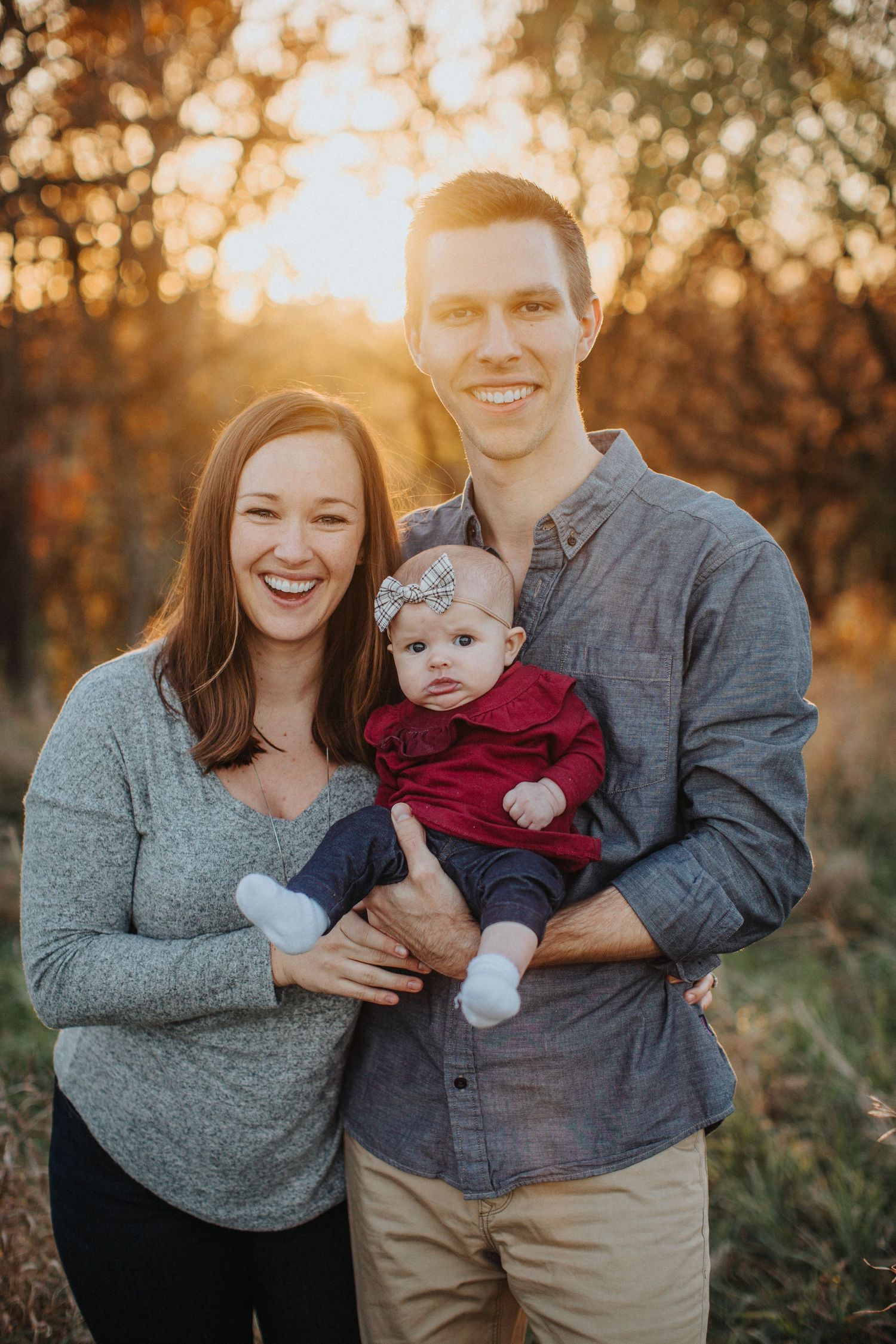 Family portrait session in a field at golden hour with warm autumn sunlight filtering through trees in the background.