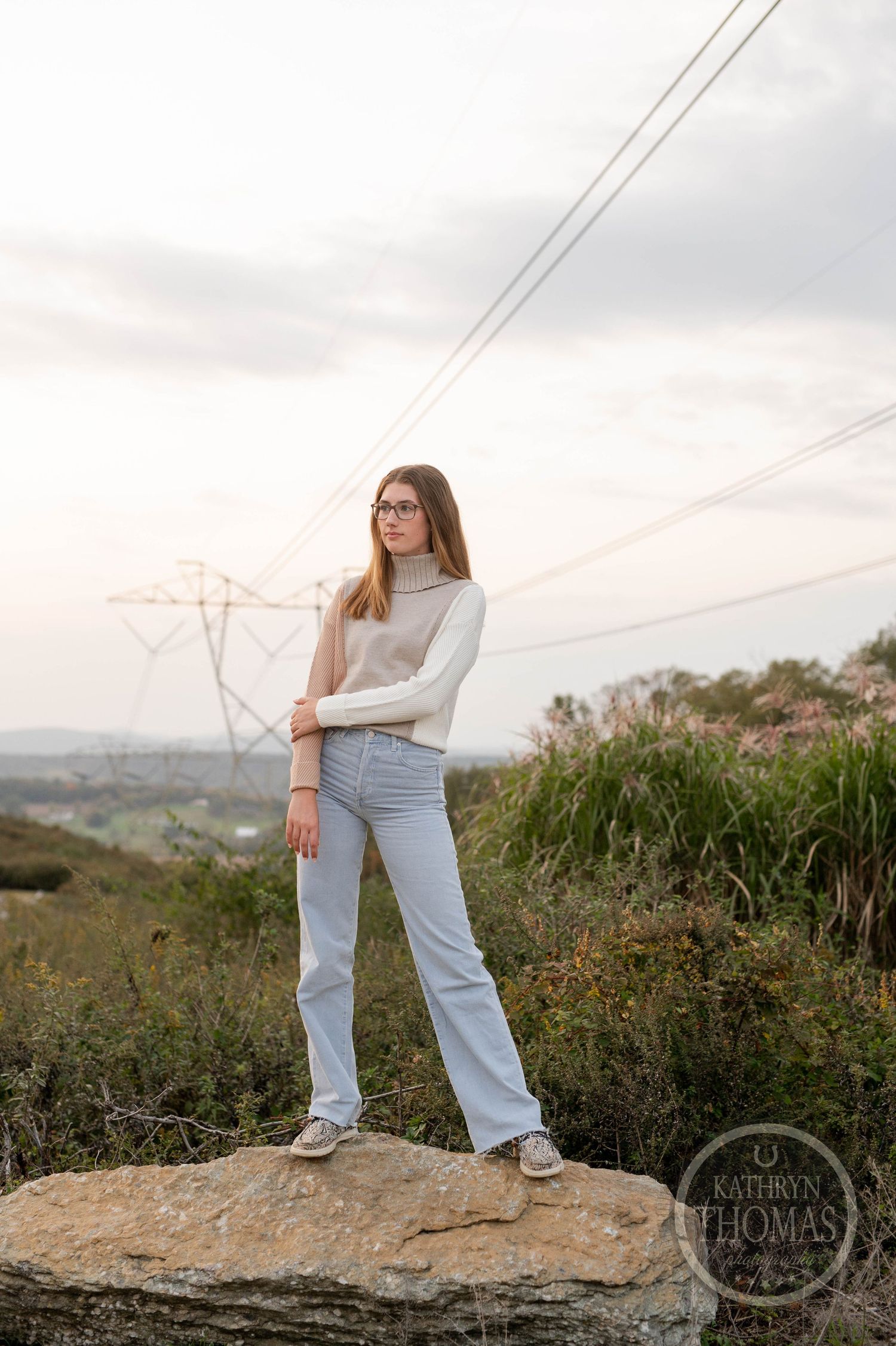 Person in glasses stands on rocky overlook wearing white sweater and light jeans with power lines in distance.