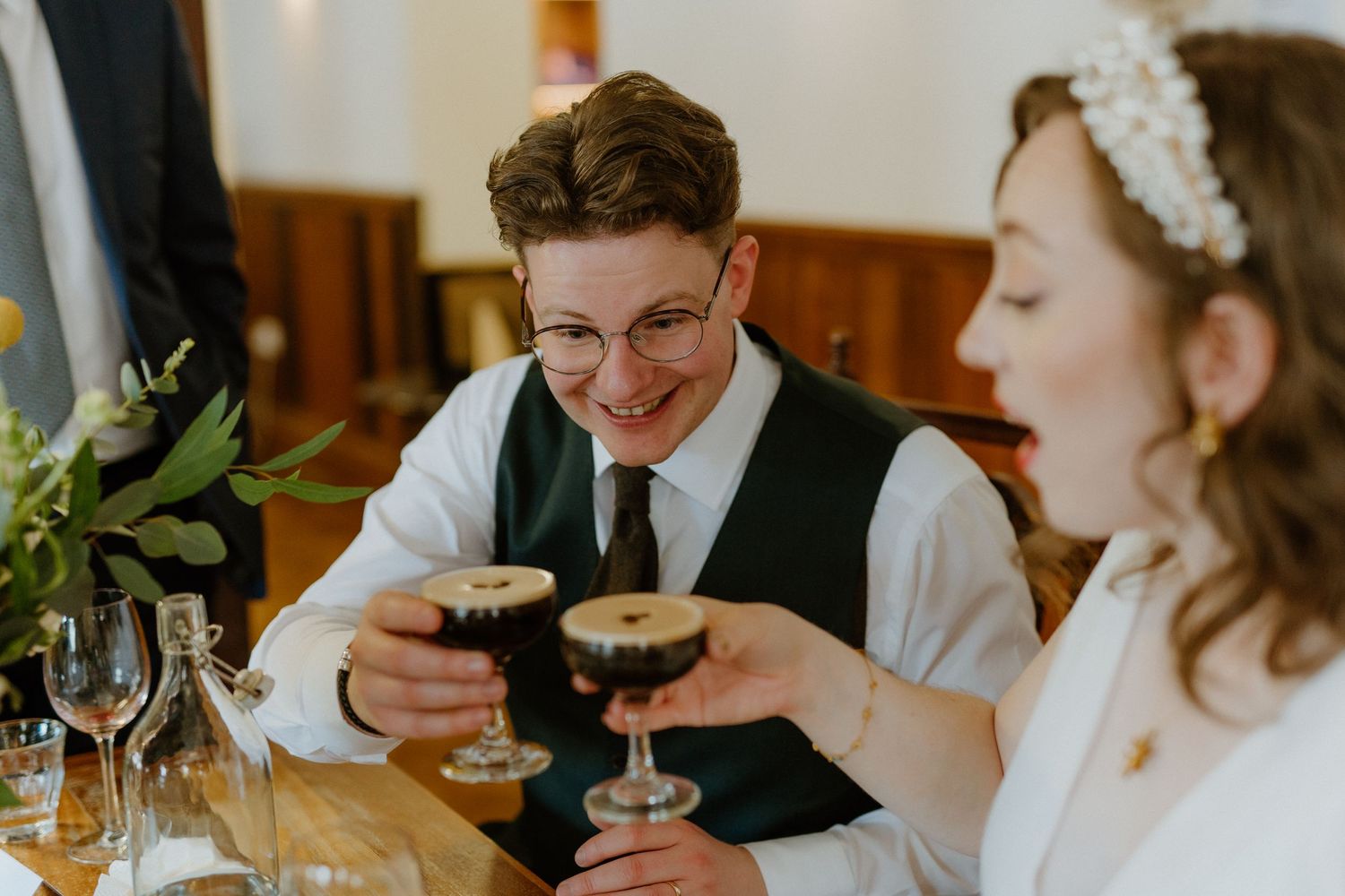 Two people in wedding attire clink glasses filled with dark cocktails while smiling at a reception table.