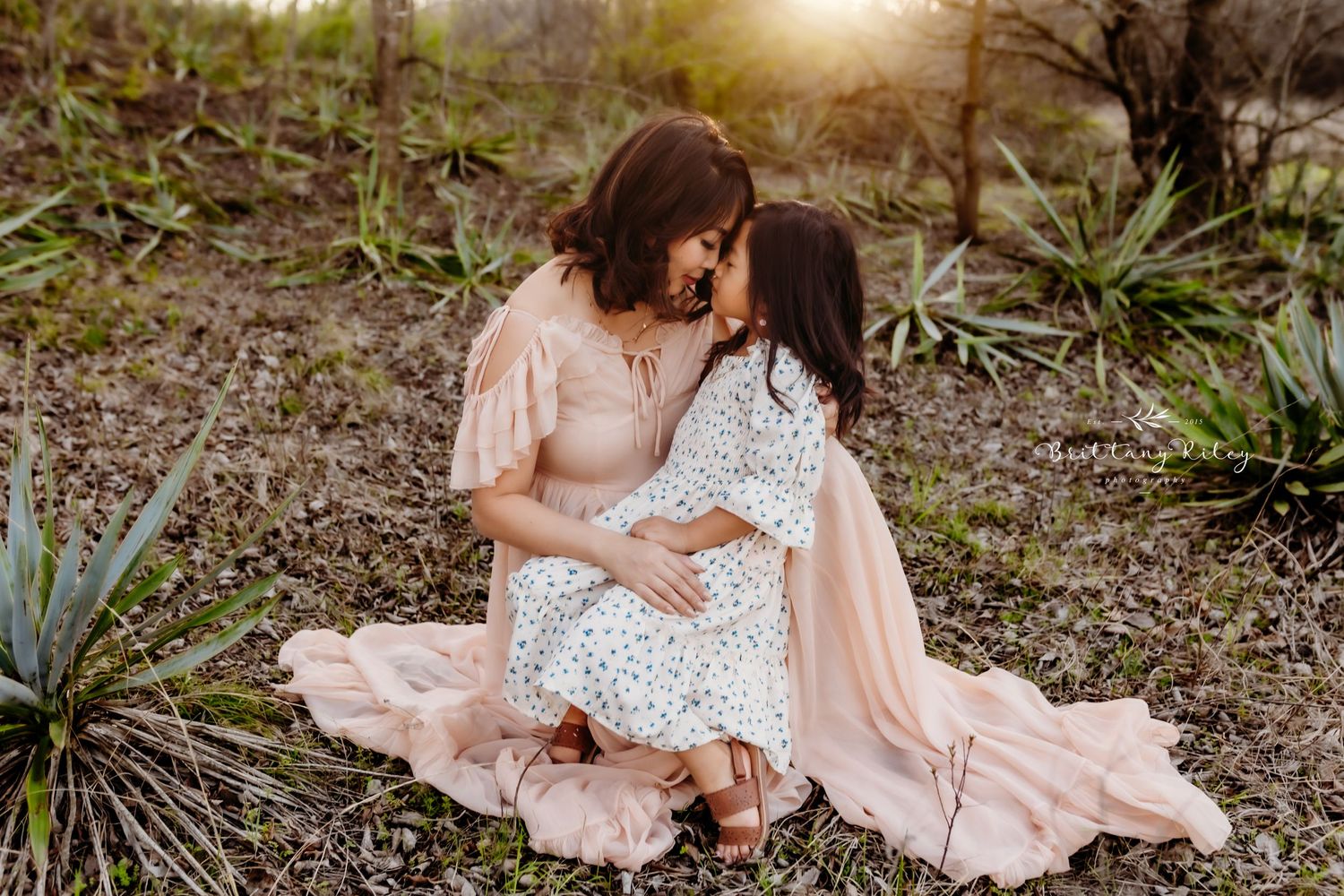 Two people in flowing maxi dresses share an intimate moment at sunset in a desert garden setting.