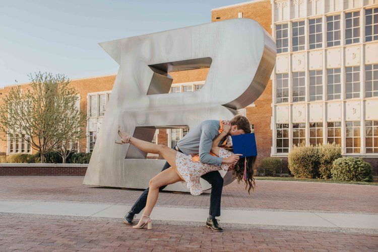 Two dancers perform an energetic dance move in front of a large letter B sculpture on a sunny day.