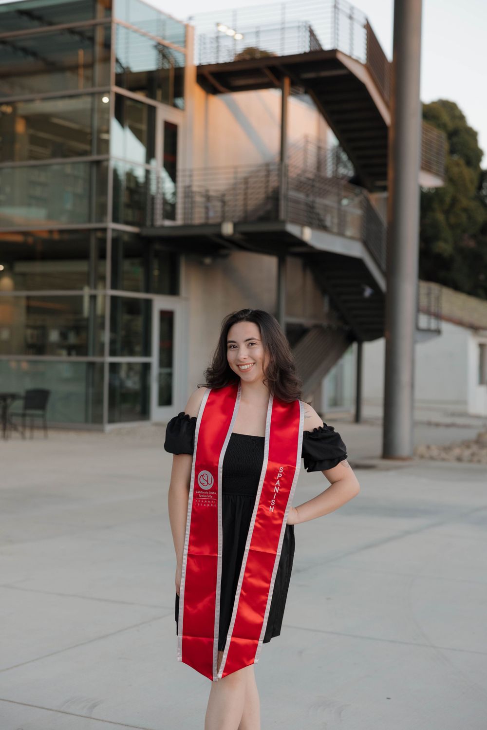 A graduate wearing a black graduation dress with red and white stoles stands outside a modern building with stairs.