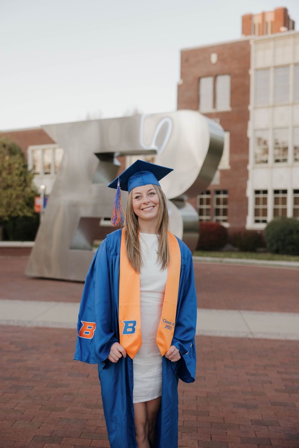 Graduate in blue cap and gown with orange stole stands smiling in front of modern campus building with brick courtyard.