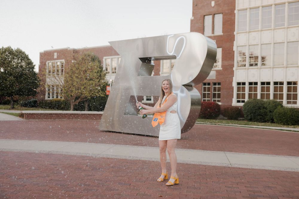 A person in a white sundress stands in front of a large metal abstract sculpture on a university campus.