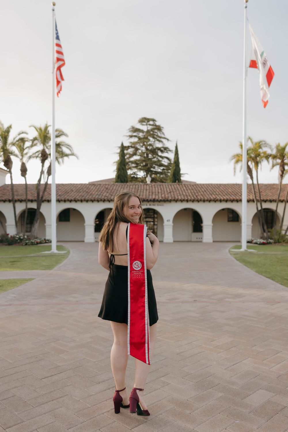 Graduate in black dress and red sash poses in front of Spanish-style building with American and Japanese flags.