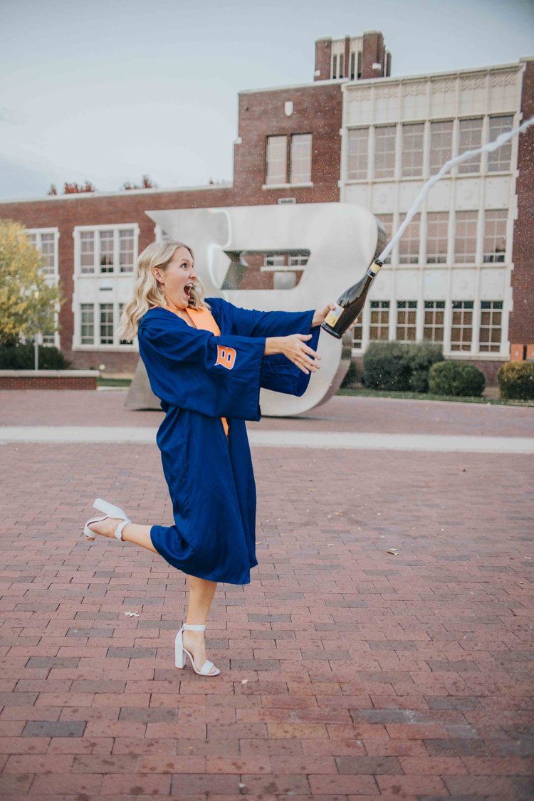Graduate in blue gown strikes joyful pose in front of campus building with modern white sculpture.