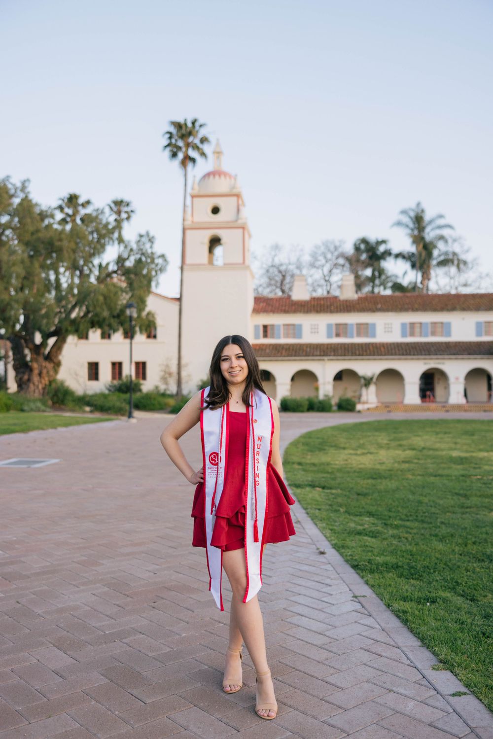 Graduate poses in a red dress and graduation stole on brick walkway in front of mission-style building with bell tower.