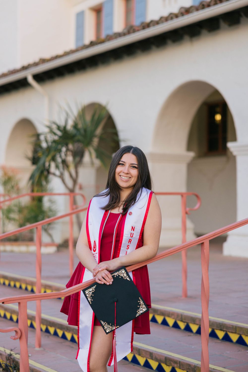 Graduate in red dress and white stole holds diploma while standing on steps with Spanish-style arches and coral railing.