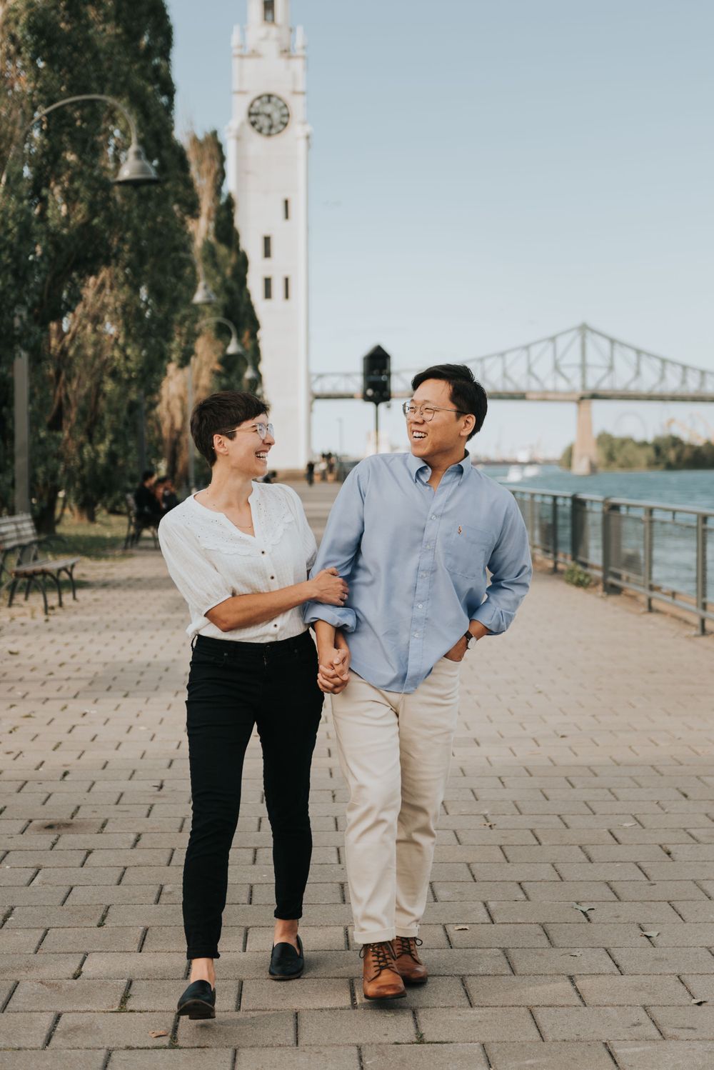 A couple walks together along the riverfront promenade with a white clock tower and bridge visible in the background.