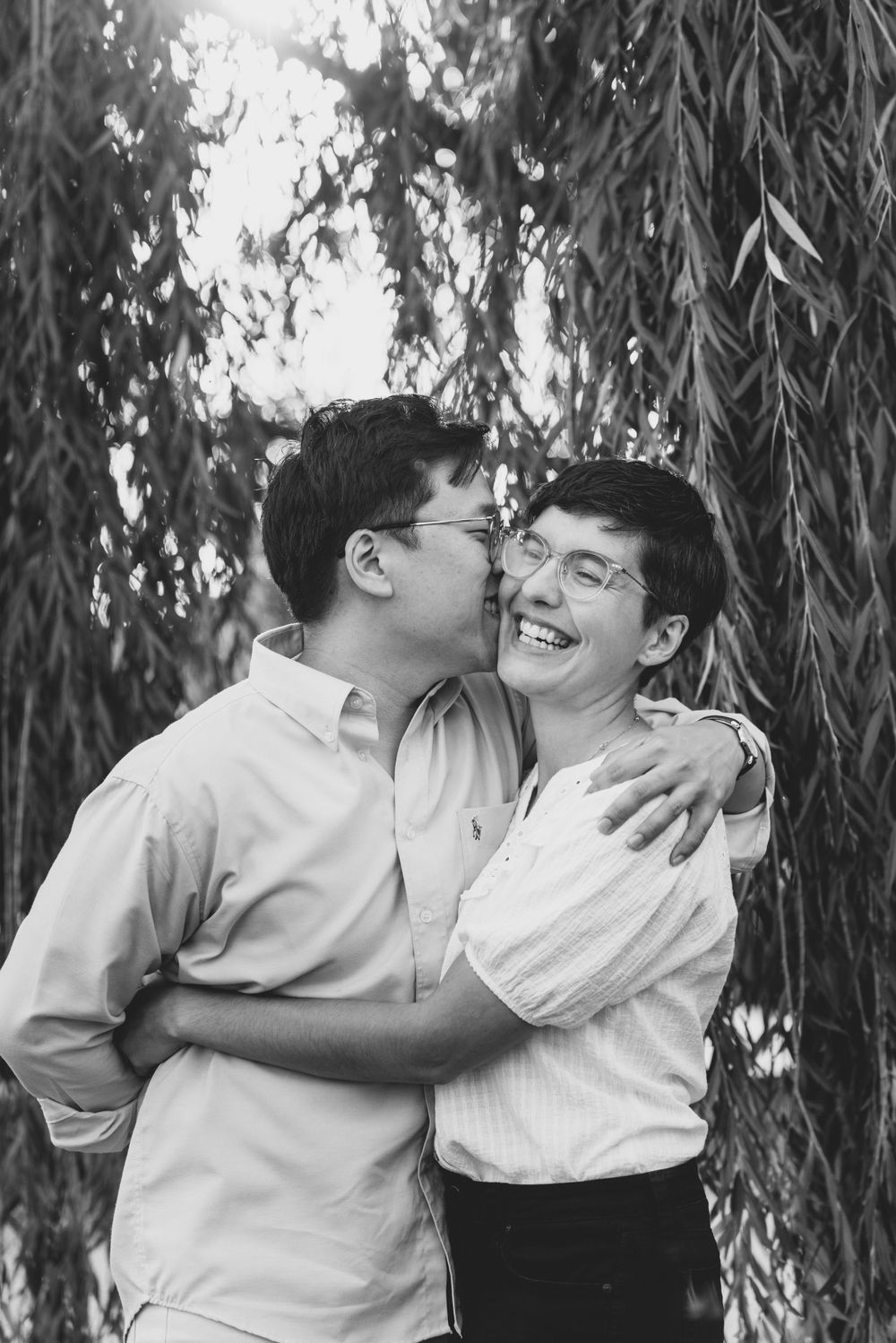 A tender moment shared between a couple embracing and laughing together under willow tree branches in black and white.