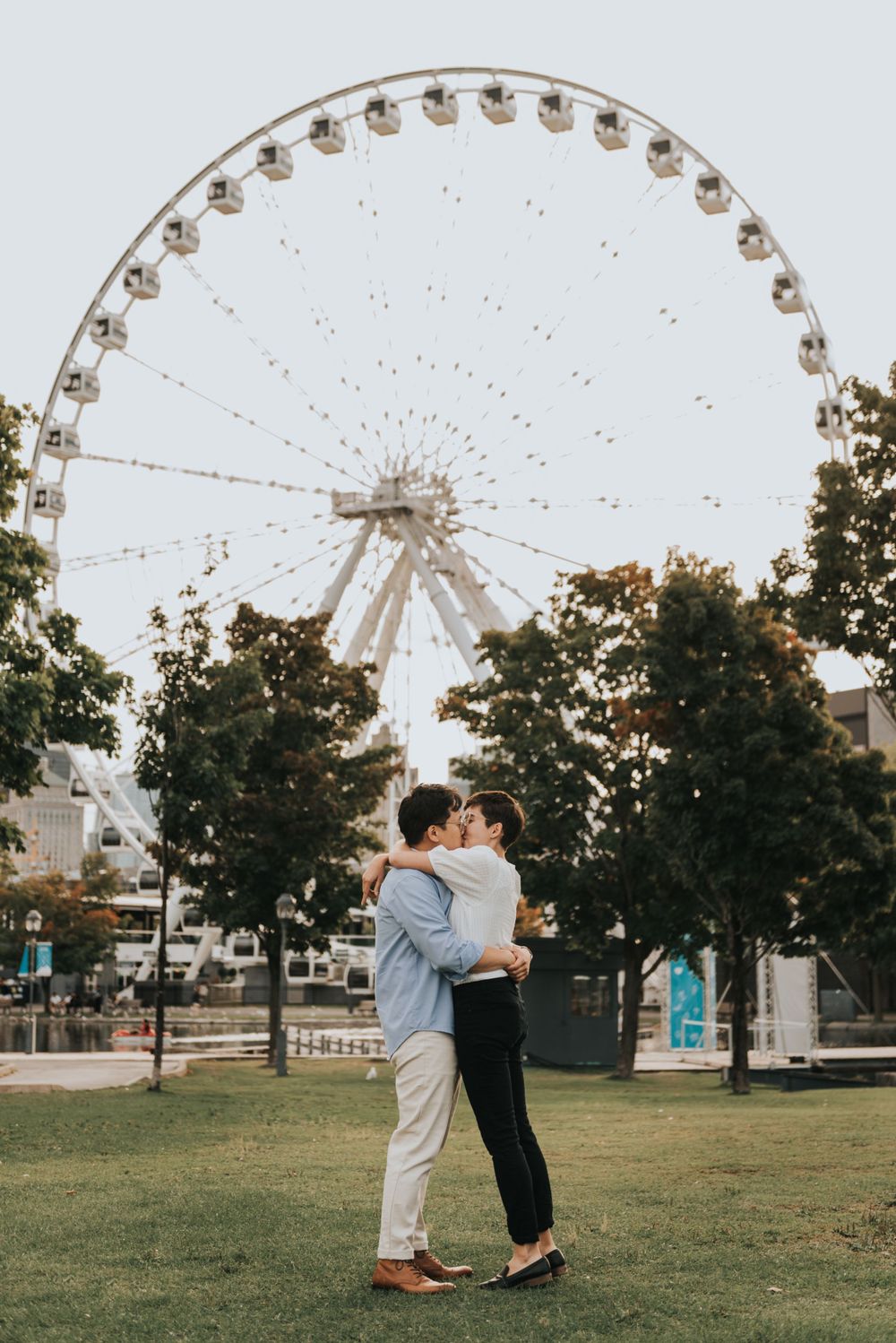 A couple embraces in front of a large Ferris wheel at sunset in a park setting.