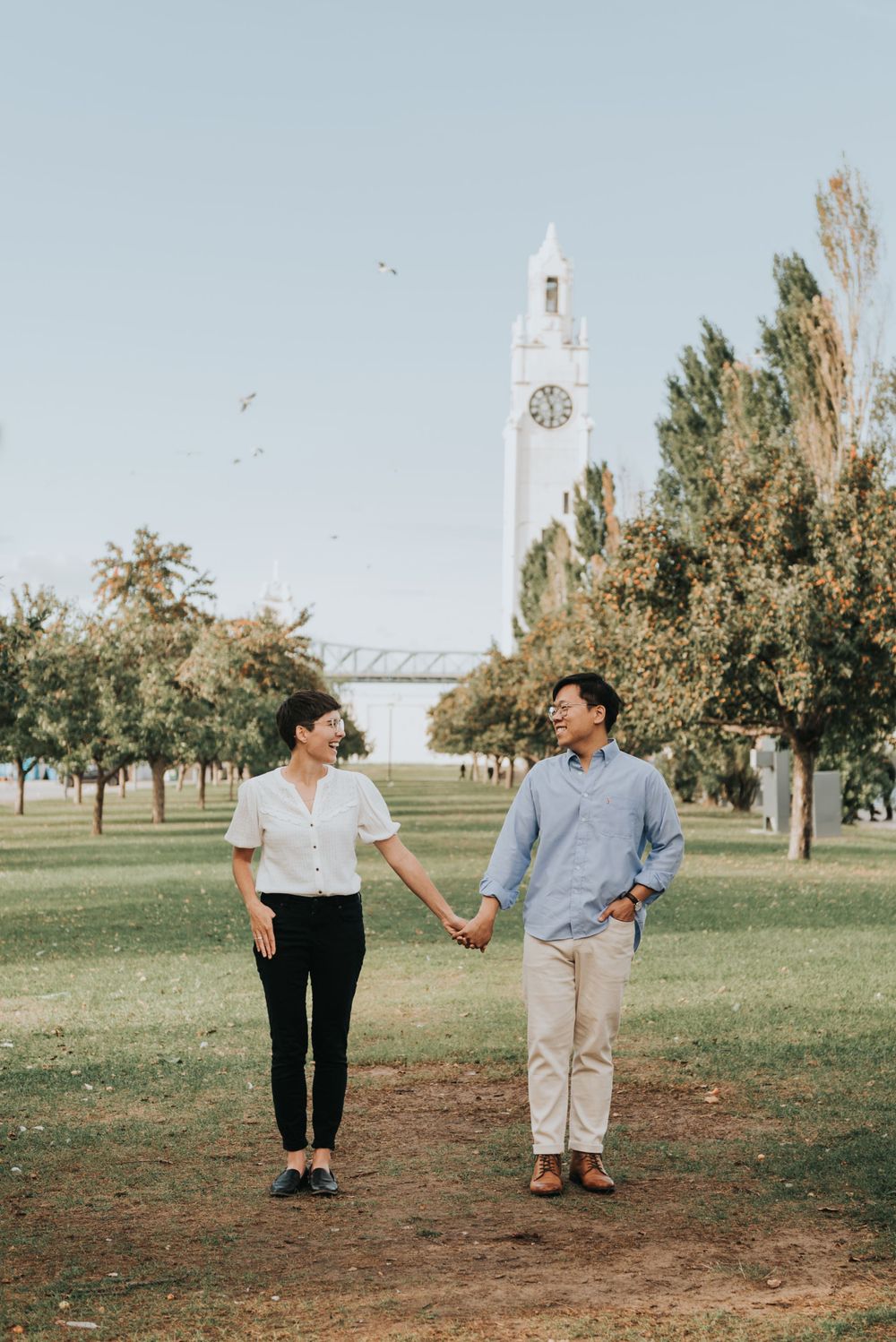 A couple walks hand in hand on a path through a park with a white clock tower visible in the background.