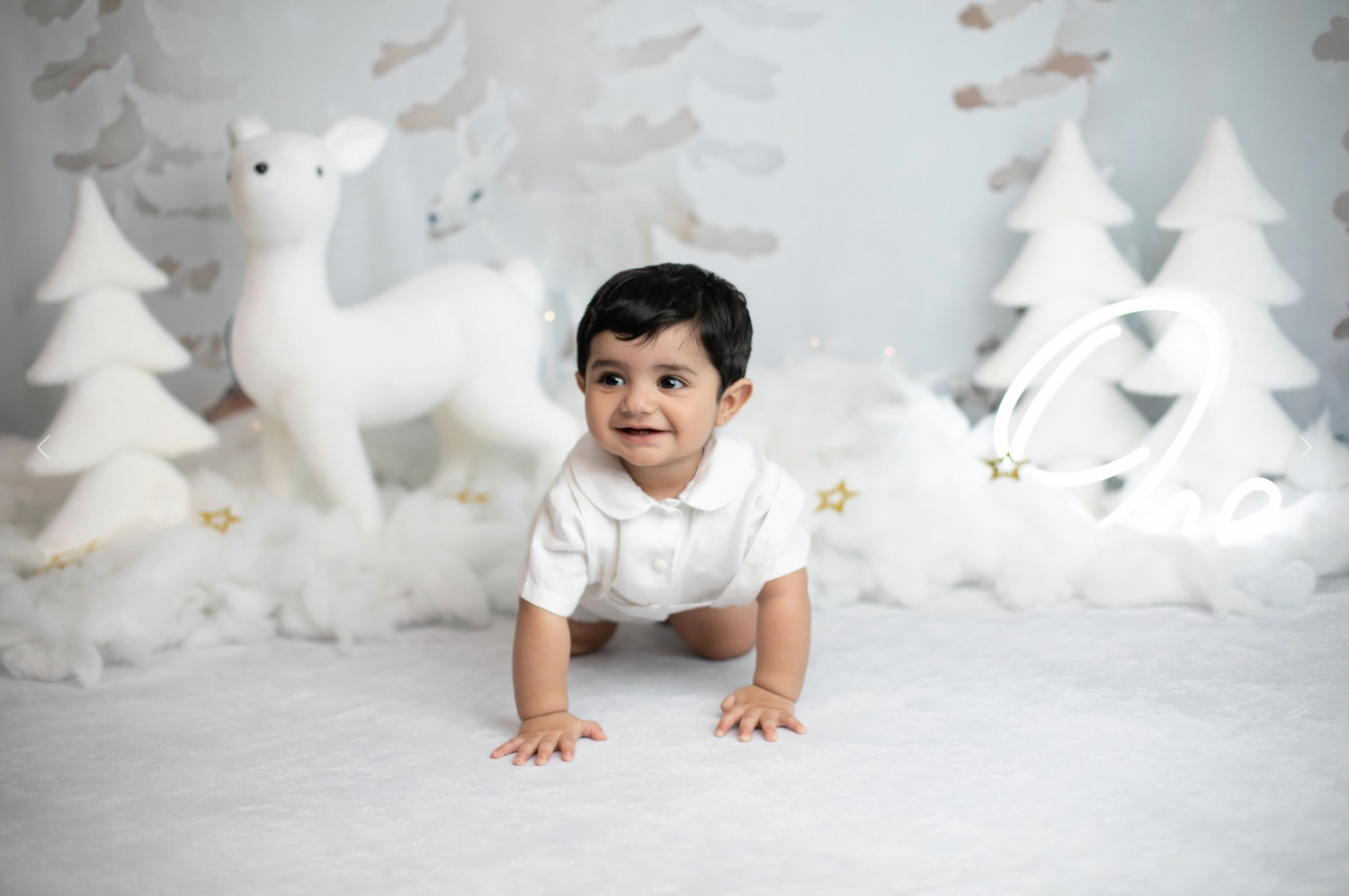 A young child in white crawls on a snowy backdrop with decorative white reindeer and trees in a Christmas-themed setting.